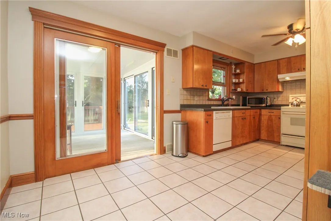 Kitchen with backsplash, white appliances, dark countertops, under cabinet range hood, and light tile patterned floors