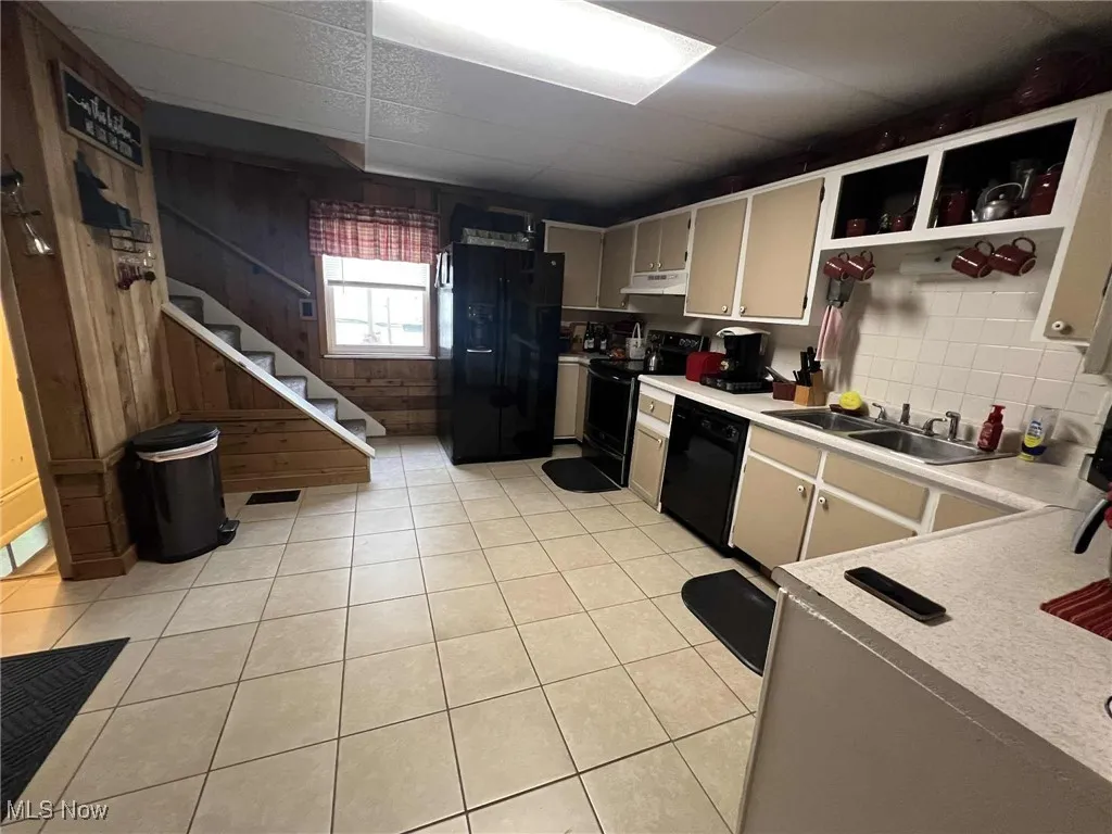 Kitchen featuring black appliances, light countertops, wooden walls, light tile patterned floors, and under cabinet range hood