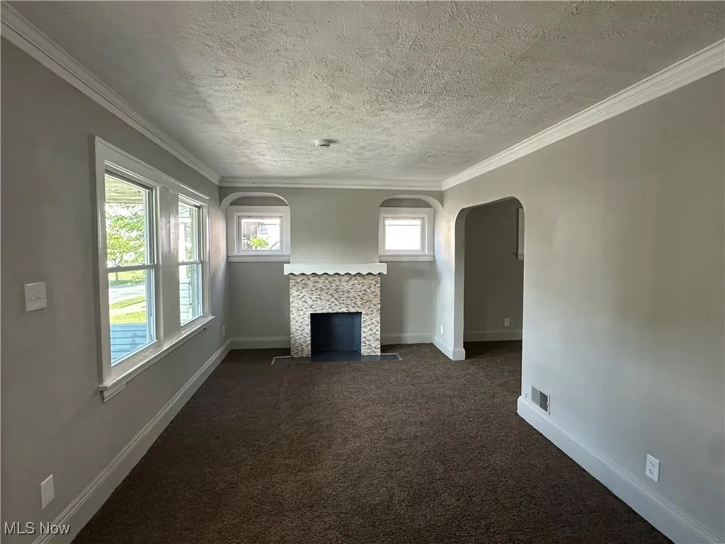 Unfurnished living room with dark colored carpet, arched walkways, a fireplace, a textured ceiling, and ornamental molding