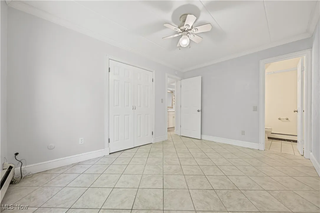 Unfurnished bedroom featuring light tile patterned flooring, ornamental molding, ensuite bath, a closet, and a baseboard heating unit