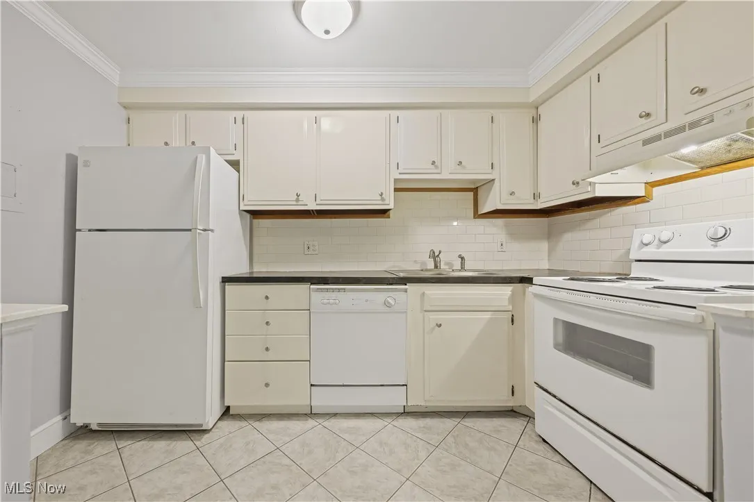 Kitchen with white appliances, ornamental molding, under cabinet range hood, backsplash, and dark countertops