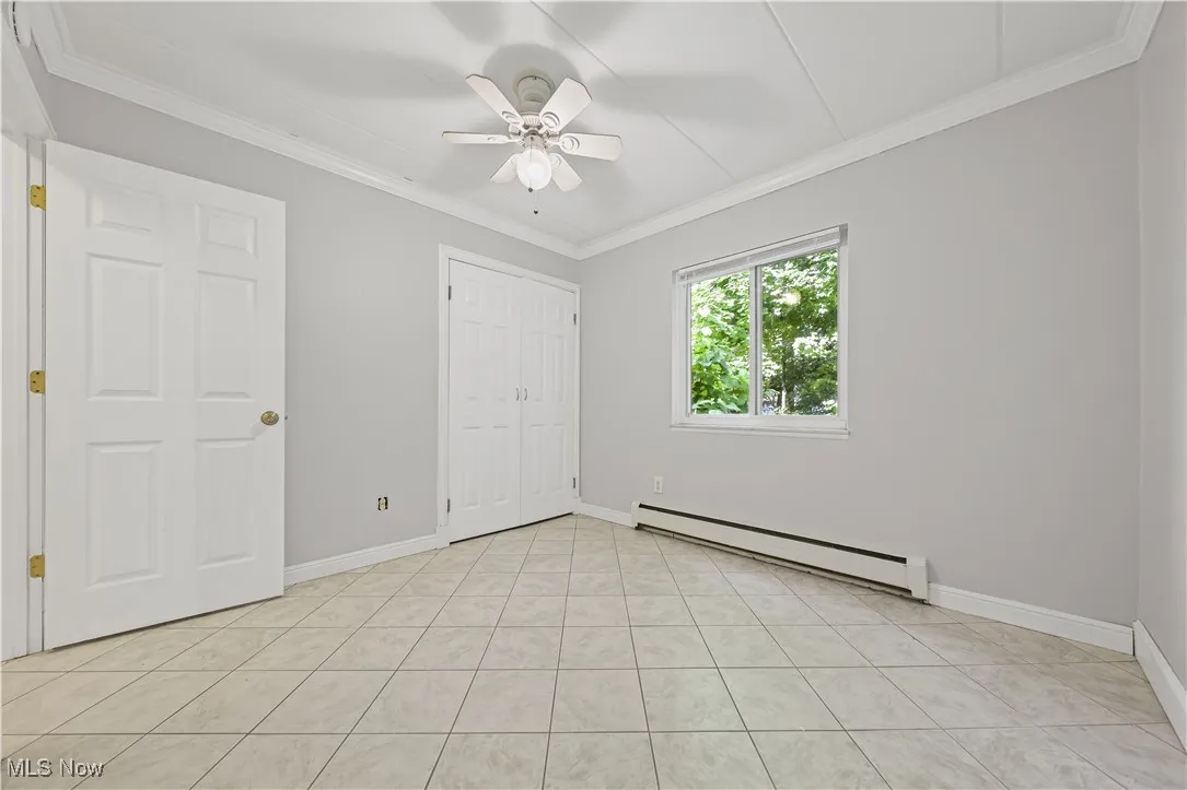 Unfurnished bedroom featuring a baseboard heating unit, crown molding, light tile patterned floors, ceiling fan, and a closet