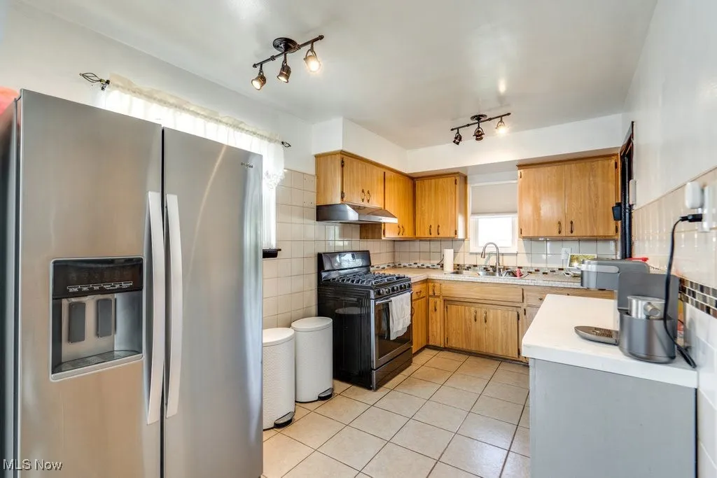 Kitchen featuring stainless steel refrigerator with ice dispenser, black gas stove, light countertops, light tile patterned floors, and under cabinet range hood