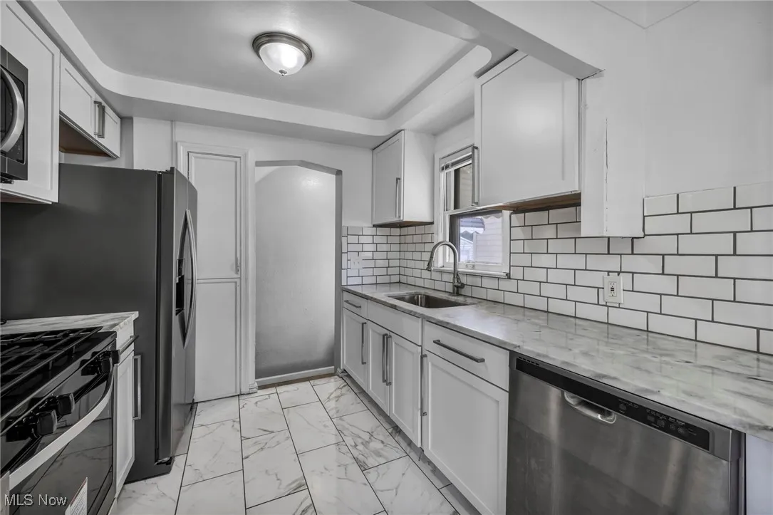 Kitchen with light marble finish floors, appliances with stainless steel finishes, tasteful backsplash, light stone countertops, and white cabinetry