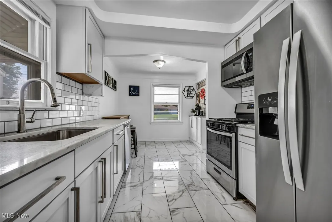 Kitchen featuring appliances with stainless steel finishes, light marble finish floors, light stone countertops, tasteful backsplash, and white cabinets