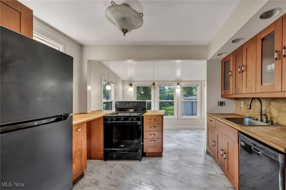 Kitchen featuring black appliances, butcher block counters, brown cabinetry, light marble finish flooring, and glass insert cabinets