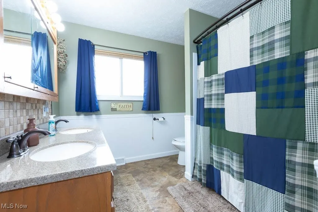 Full bath featuring wainscoting, double vanity, a textured ceiling, curtained shower, and stone finish flooring