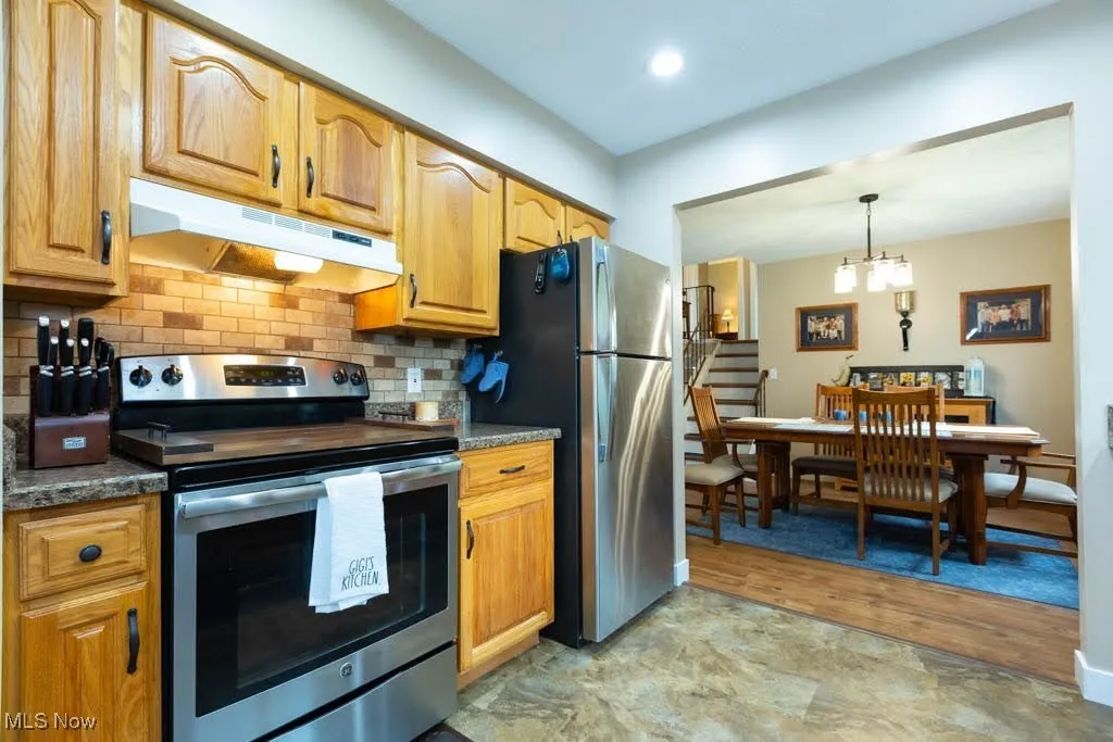 Kitchen with stainless steel appliances, under cabinet range hood, decorative backsplash, dark countertops, and a chandelier