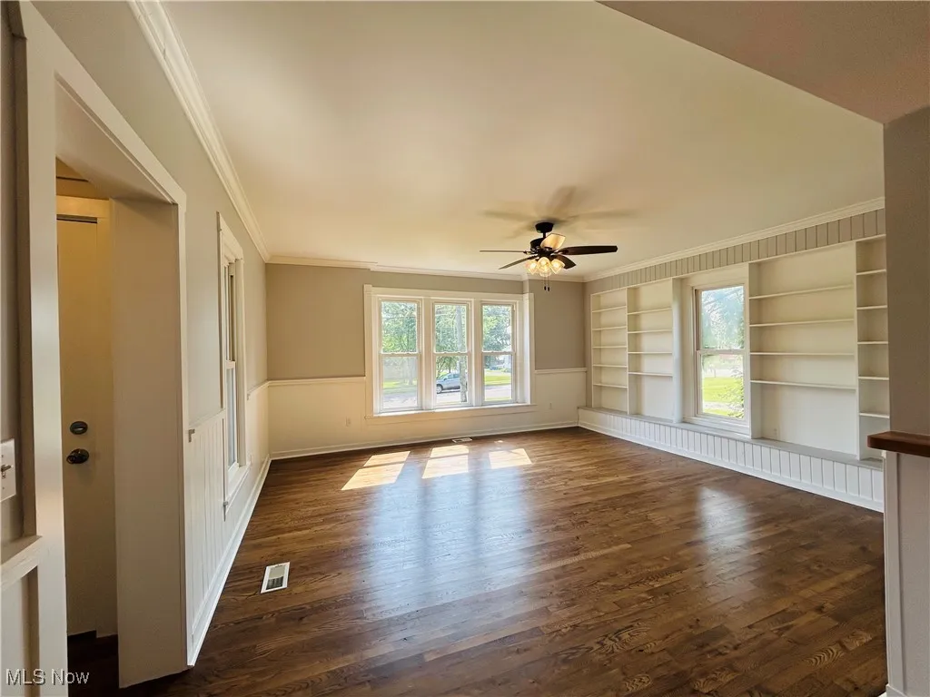Unfurnished room featuring built in features, crown molding, dark wood-style flooring, a ceiling fan, and a wainscoted wall