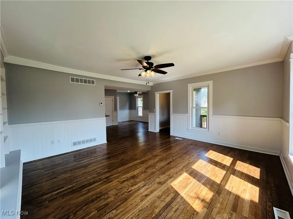 Empty room featuring a wainscoted wall, a ceiling fan, crown molding, and dark wood-style flooring