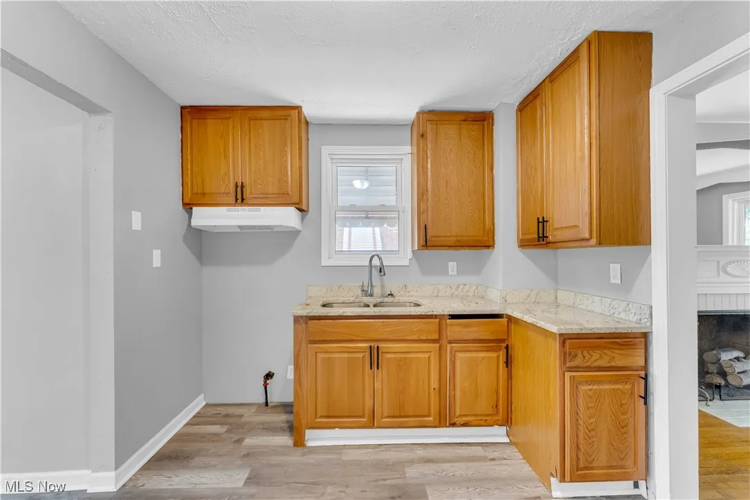 Kitchen with light wood-type flooring, light stone countertops, brown cabinetry, a textured ceiling, and under cabinet range hood