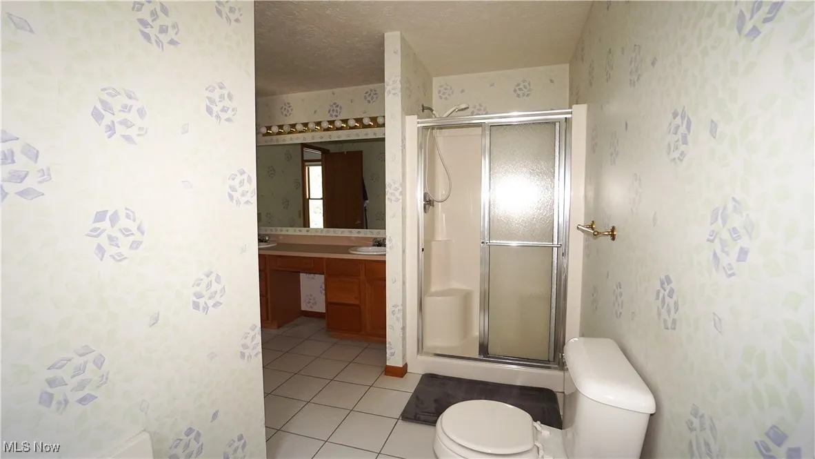 Primary bath featuring a textured ceiling, wallpapered walls, a stall shower, tile patterned flooring, and double vanity