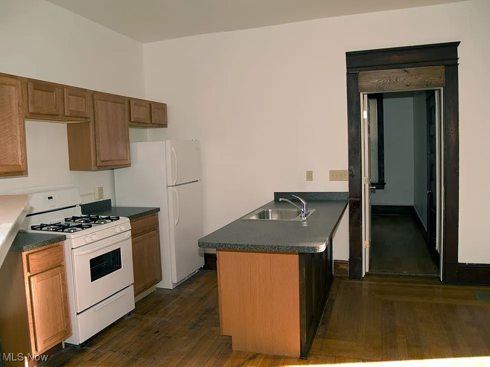 Kitchen featuring white appliances, dark wood-style flooring, dark countertops, a peninsula, and brown cabinetry