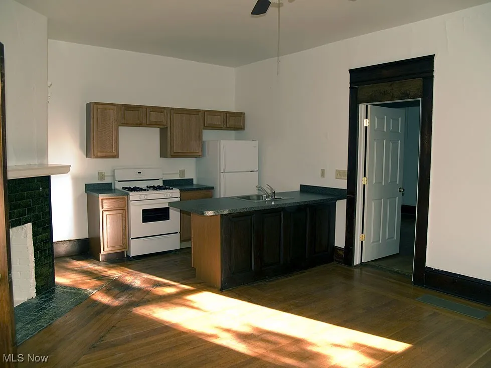 Kitchen with a peninsula, white appliances, dark wood-style floors, a brick fireplace, and dark countertops