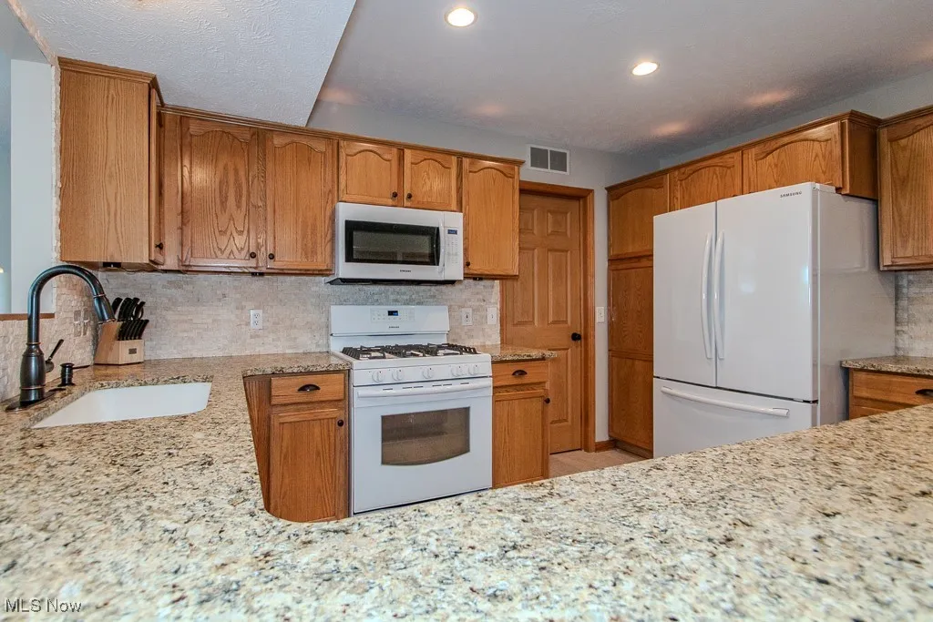 Kitchen with white appliances, tasteful backsplash, light stone counters, recessed lighting, and brown cabinetry