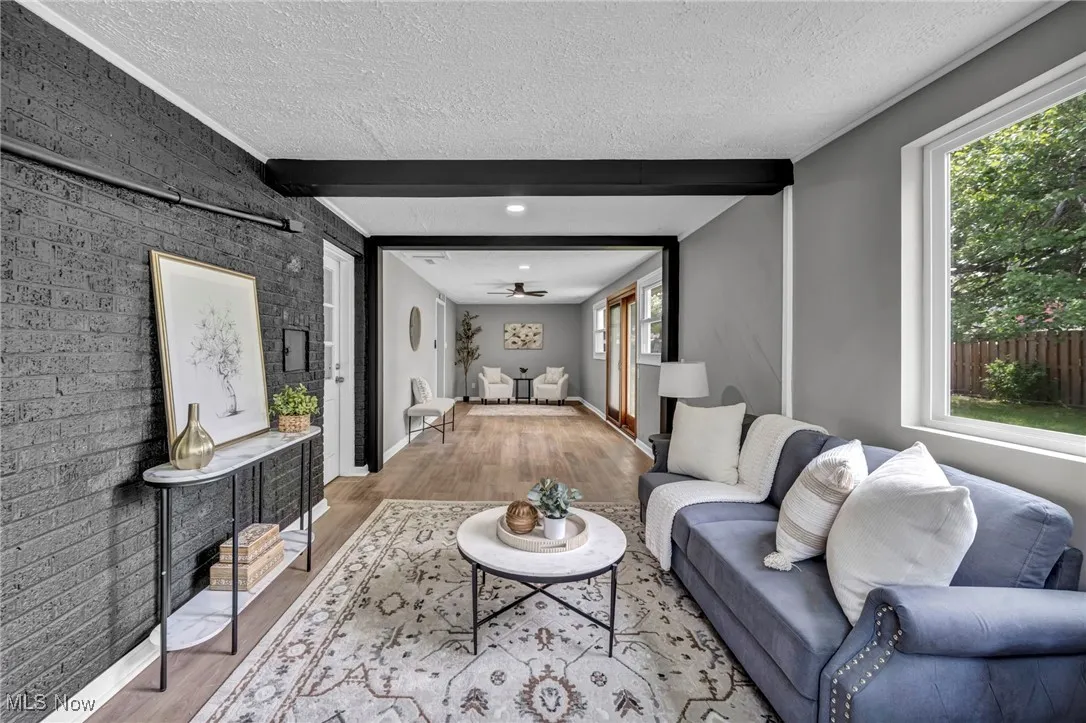 Living room with beamed ceiling, wood finished floors, a textured ceiling, brick wall, and a ceiling fan
