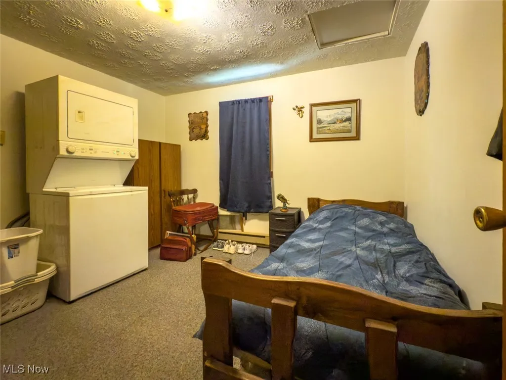 Bedroom featuring stacked washer / drying machine, a textured ceiling, attic access, carpet, and baseboard heating