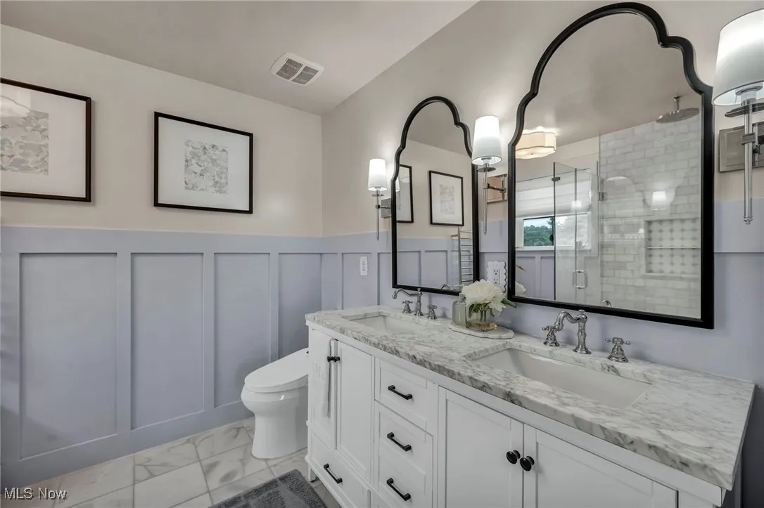 Full bath with marble finish floors, a shower stall, double vanity, wainscoting, and a decorative wall