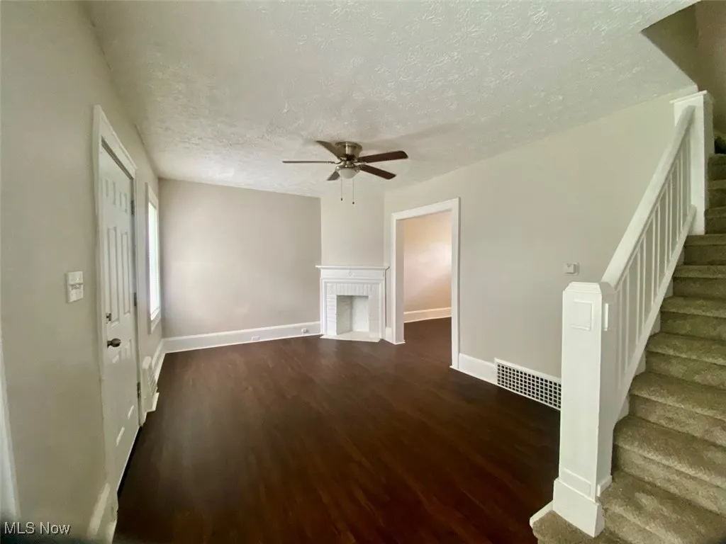 Unfurnished living room with stairs, dark wood-type flooring, a textured ceiling, a brick fireplace, and a ceiling fan