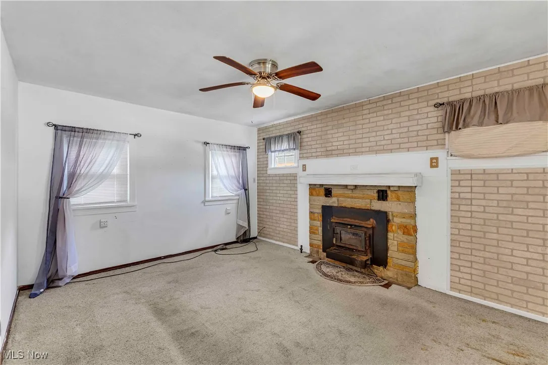 Unfurnished living room with brick wall, carpet flooring, a wood stove, and ceiling fan