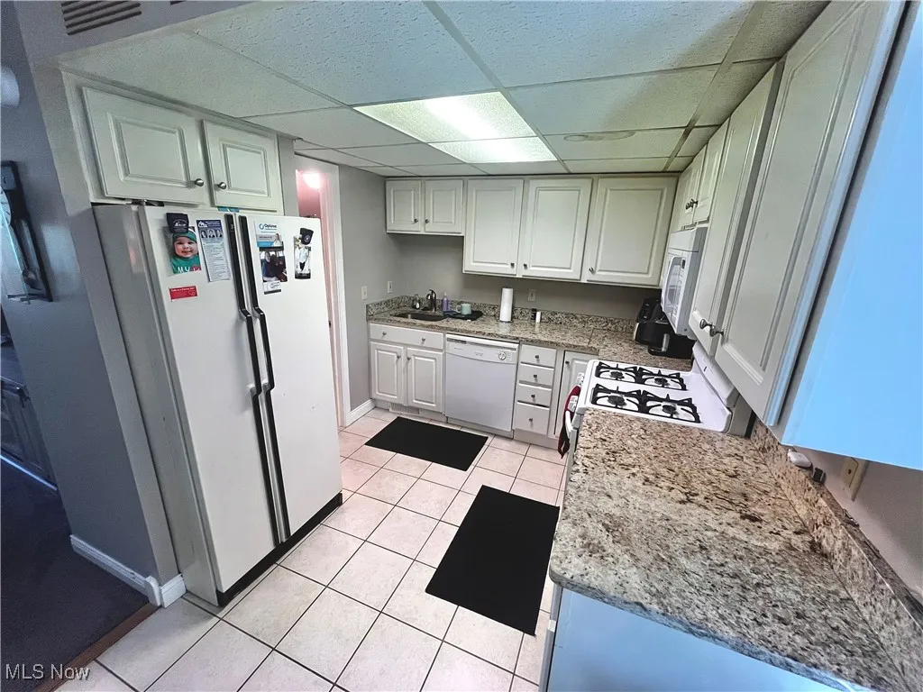 Kitchen featuring a drop ceiling, white appliances, light tile patterned floors, light stone countertops, and white cabinets