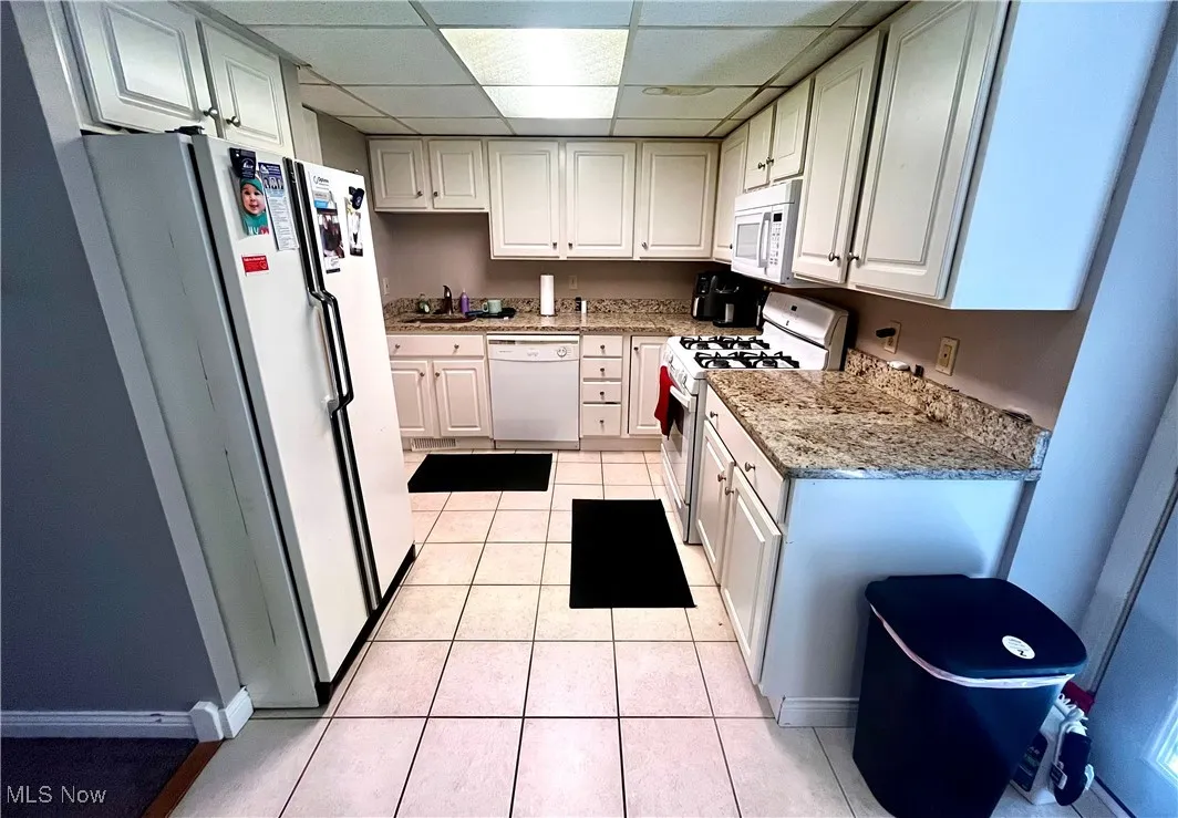 Kitchen featuring a drop ceiling, white appliances, light tile patterned floors, light stone counters, and white cabinets