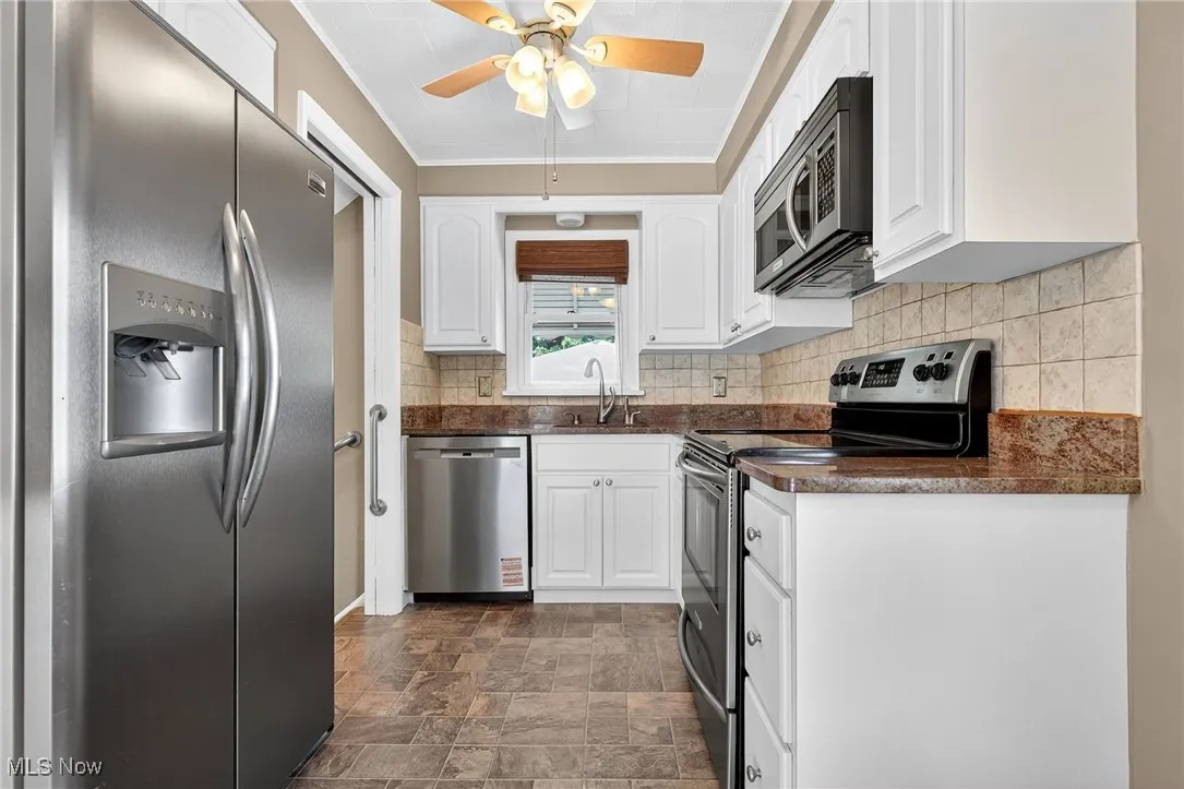 Kitchen with stainless steel appliances, dark countertops, stone finish floors, white cabinetry, and ornamental molding