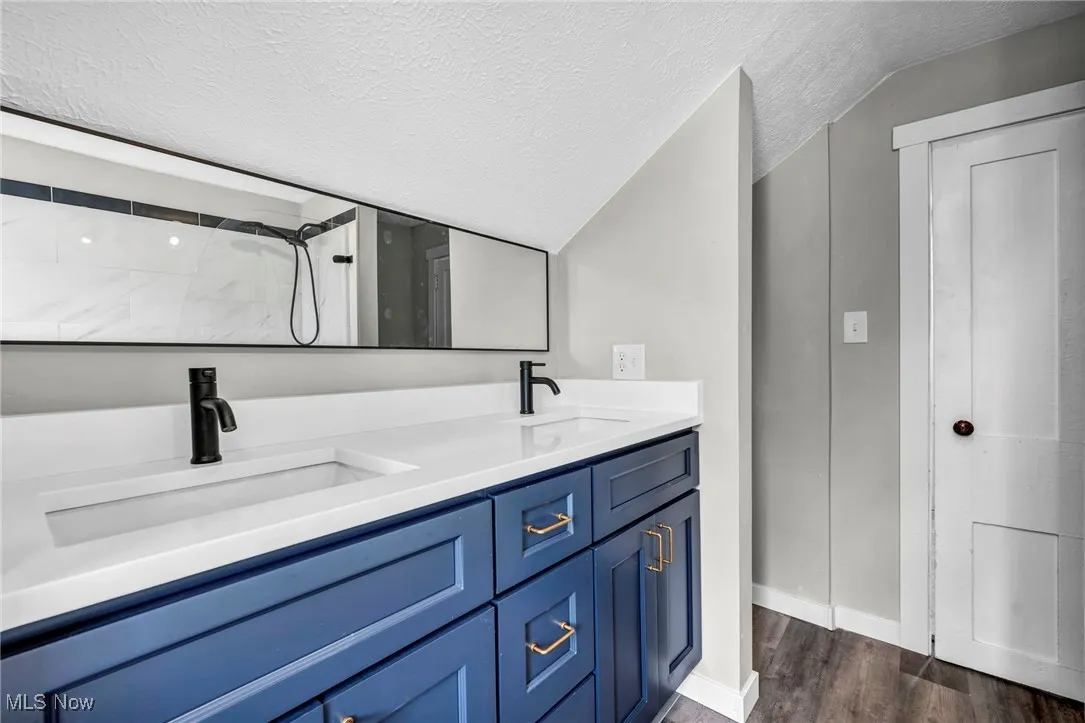 Bathroom featuring a textured ceiling, tiled shower, wood finished floors, double vanity, and lofted ceiling