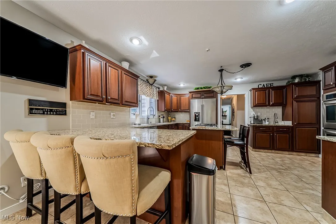 Kitchen featuring light tile patterned floors, a peninsula, tasteful backsplash, a kitchen bar, and light granite countertops.  Wall mounted TV stays