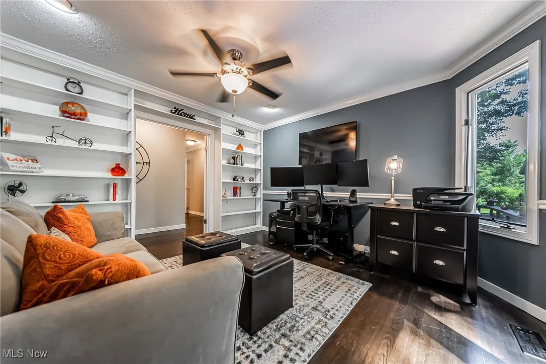 Living room with a desk, dark wood-style floors, crown molding, built in shelves, and ceiling fan