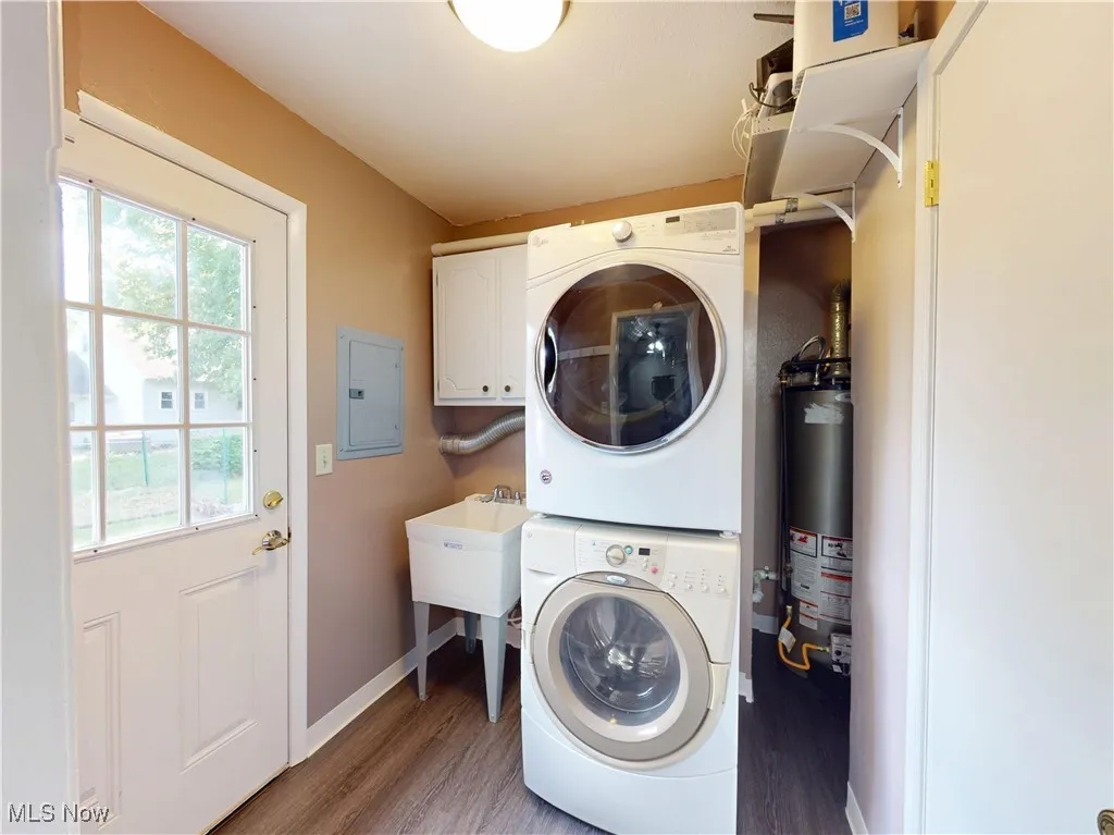 Laundry room featuring stacked washer / drying machine, cabinet space, light wood-type flooring, electric panel, and water heater