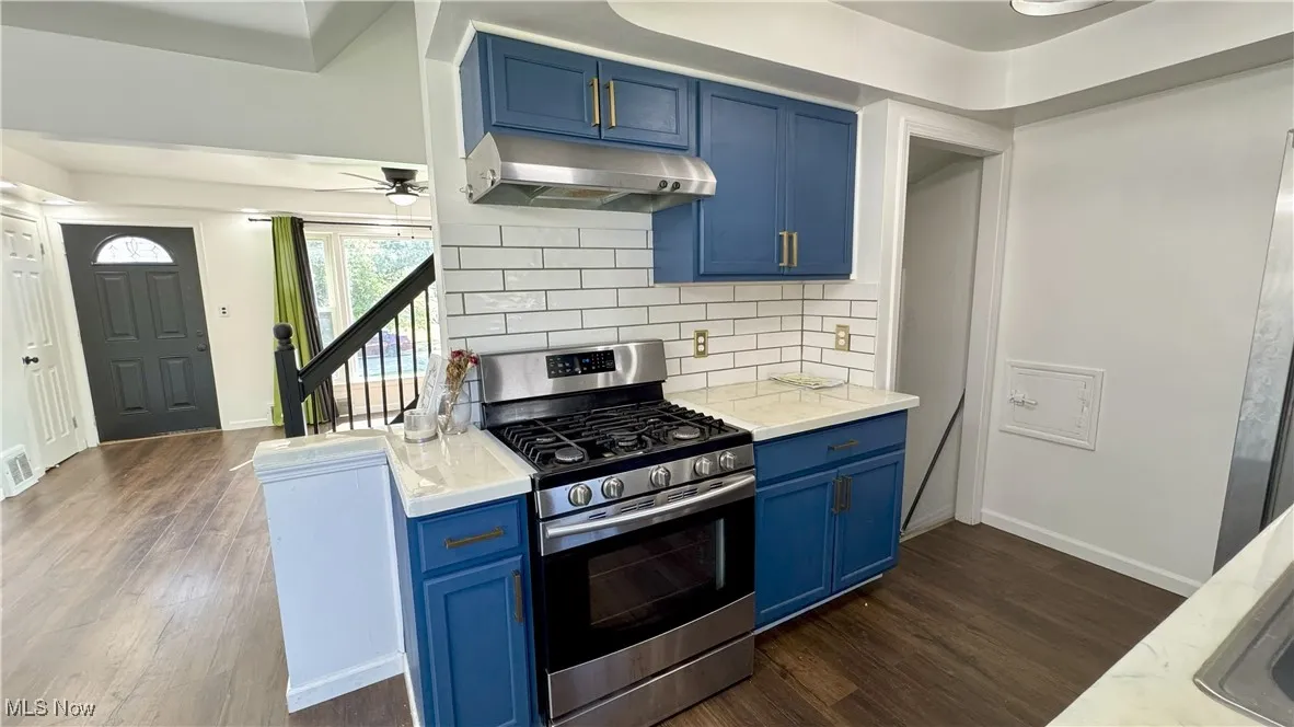 Kitchen with blue cabinets, gas stove, under cabinet range hood, tasteful backsplash, and dark wood-style floors