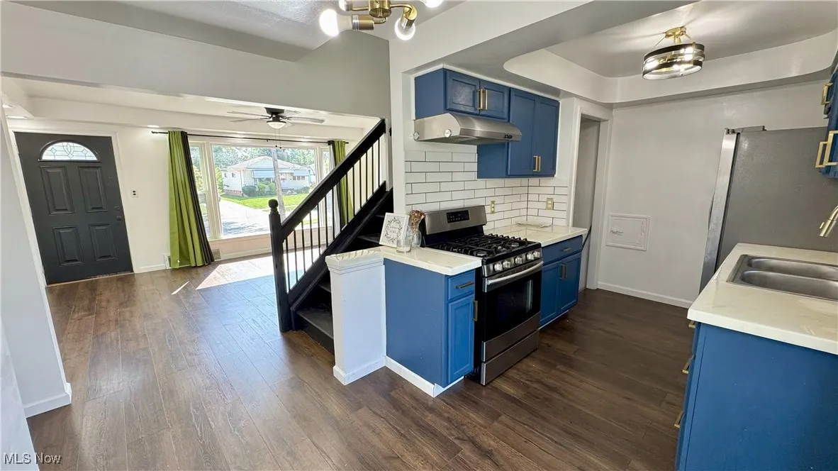 Kitchen featuring blue cabinets, backsplash, stainless steel appliances, dark wood-style flooring, and under cabinet range hood