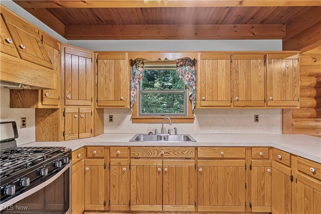 Kitchen featuring stainless steel gas stove, custom exhaust hood, light countertops, brown cabinets, and a wood ceiling with exposed beams