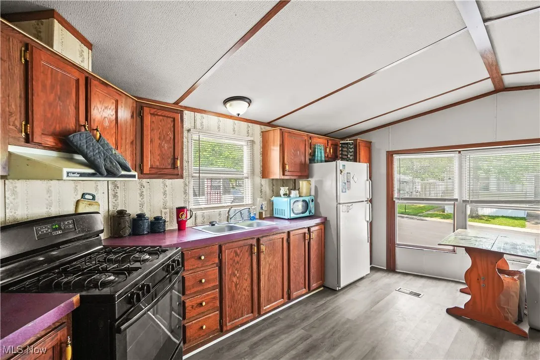 Kitchen featuring black gas range, vaulted ceiling, under cabinet range hood, dark wood finished floors, and freestanding refrigerator