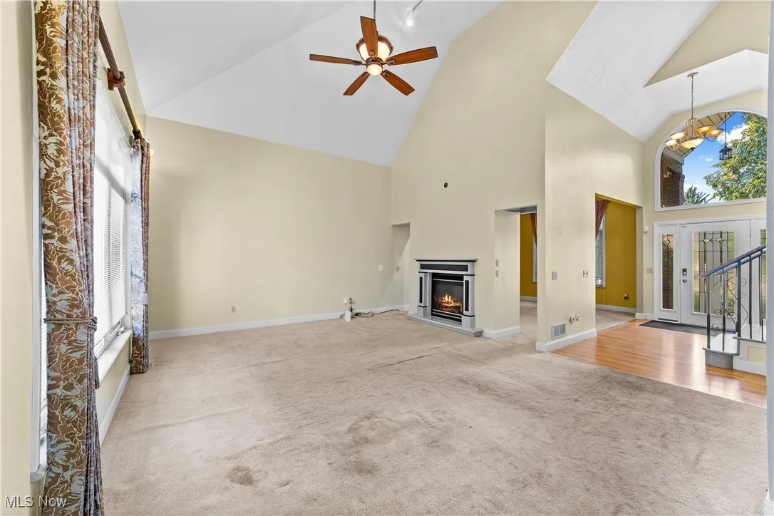 Unfurnished living room featuring high vaulted ceiling, a lit fireplace, light colored carpet, a ceiling fan, and stairs