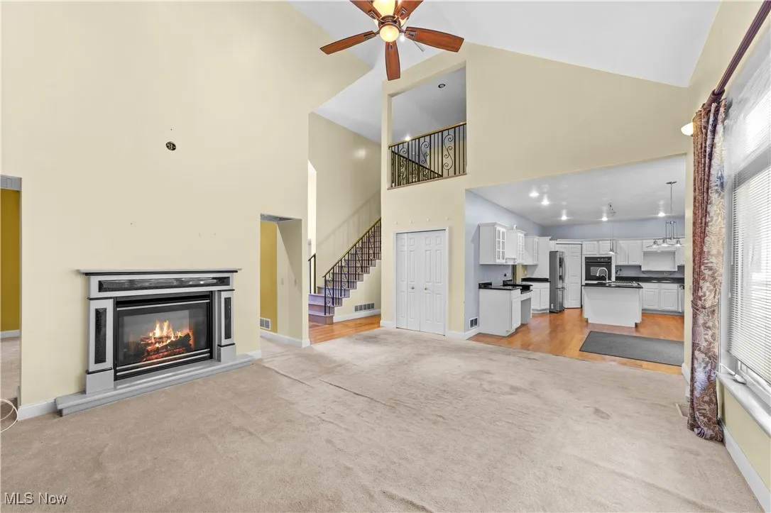 Unfurnished living room featuring a glass covered fireplace, light colored carpet, high vaulted ceiling, stairs, and ceiling fan