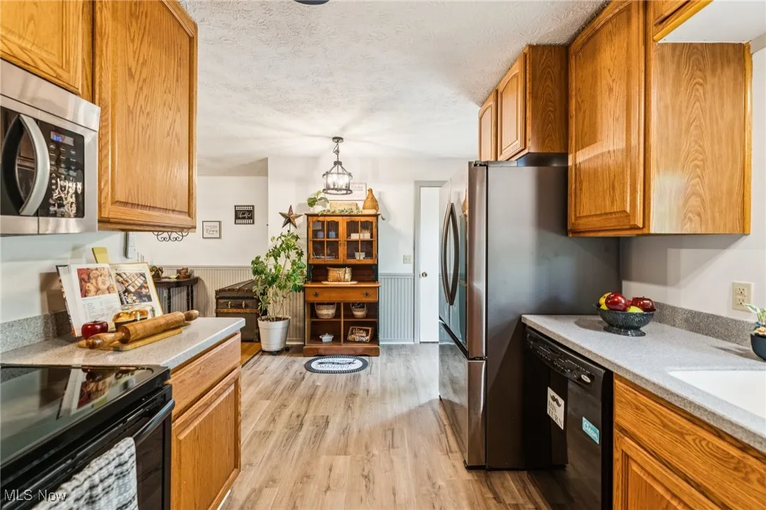 Kitchen with a wainscoted wall, black appliances, a textured ceiling, brown cabinetry, and light wood-style flooring