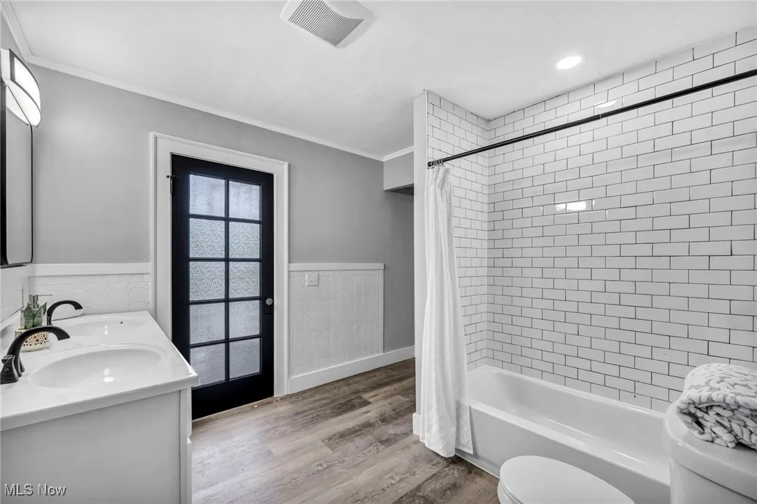 Bathroom with wainscoting, shower / tub combo with curtain, light wood-style flooring, ornamental molding, and tile walls