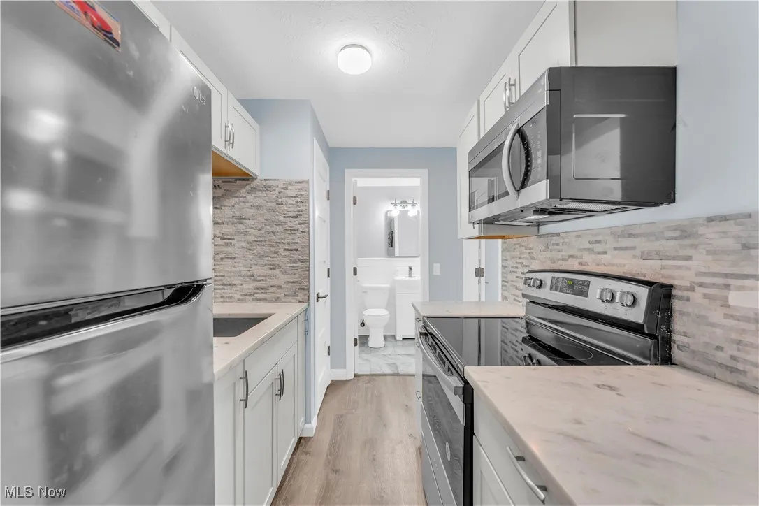 Kitchen with stainless steel appliances, white cabinetry, light wood-type flooring, tasteful backsplash, and light stone counters