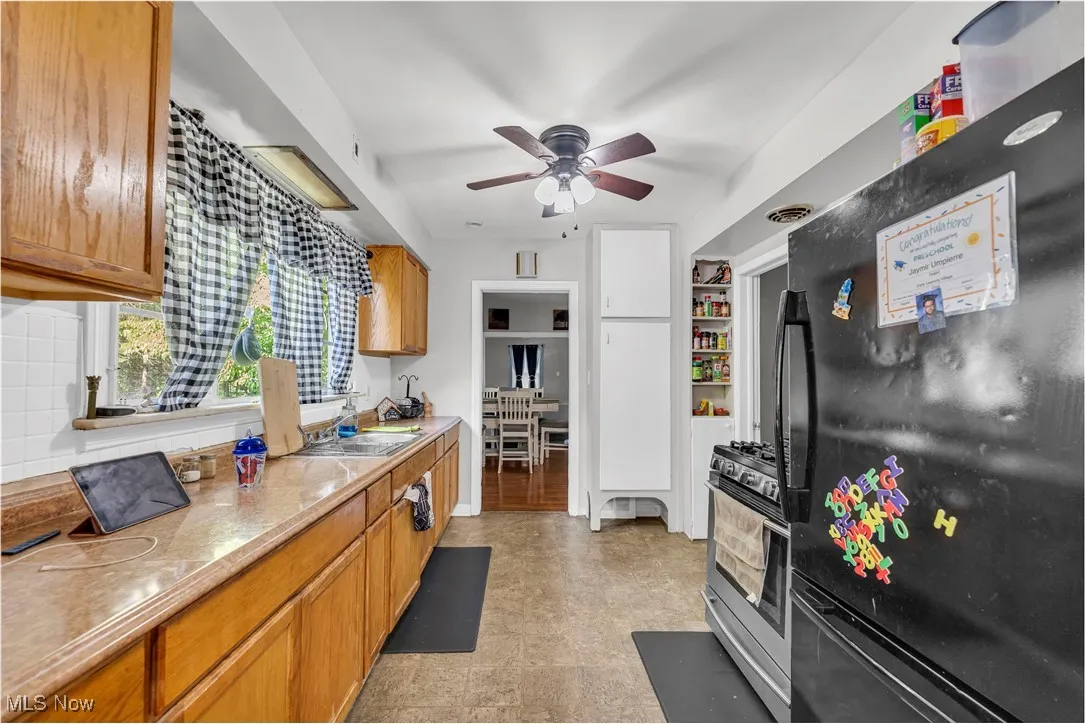 Kitchen with freestanding refrigerator, gas stove, brown cabinetry, light countertops, and ceiling fan