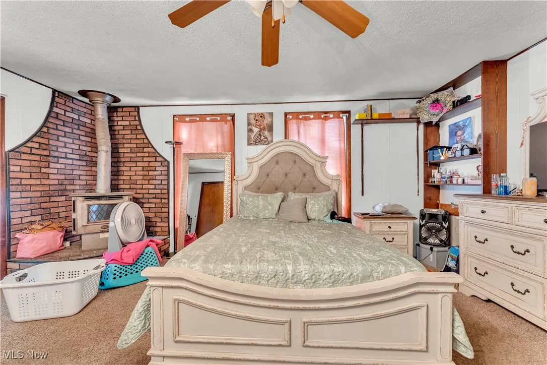 Bedroom with a textured ceiling, light carpet, a wood stove, a ceiling fan, and brick wall