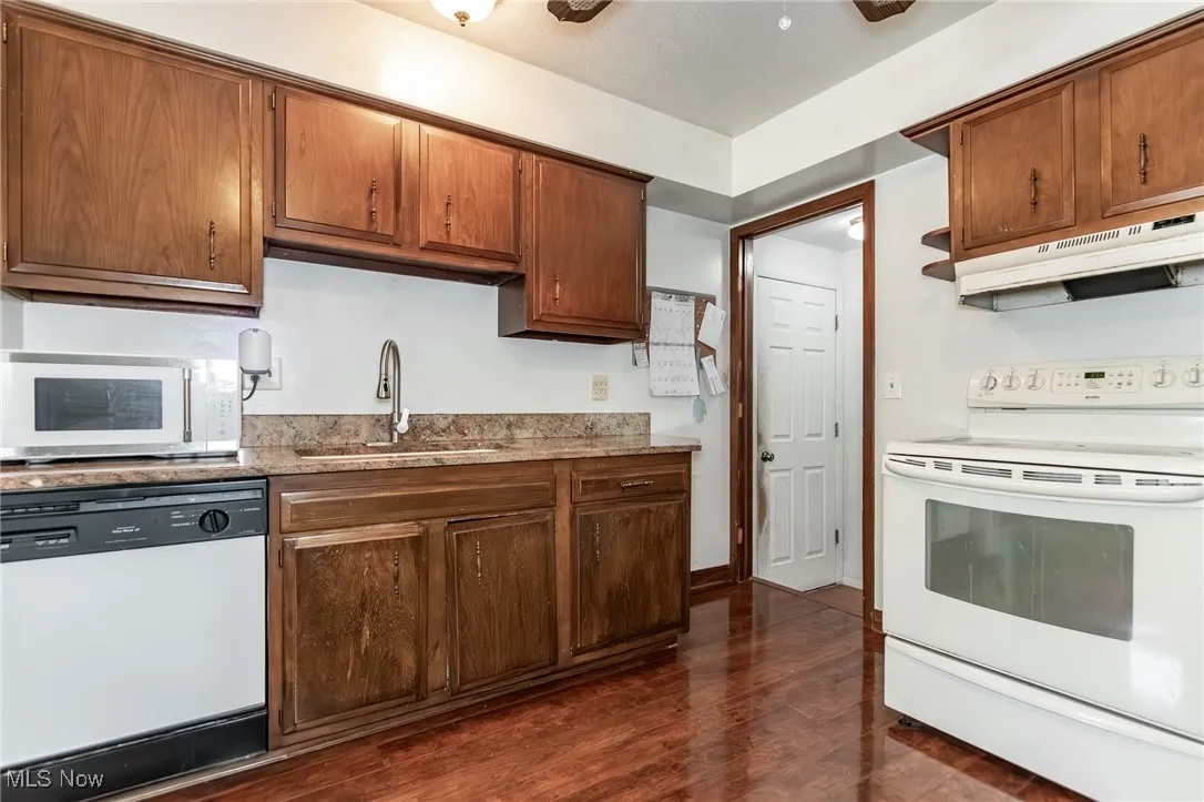 Kitchen with white appliances, under cabinet range hood, dark wood-style floors, light stone counters, and brown cabinetry