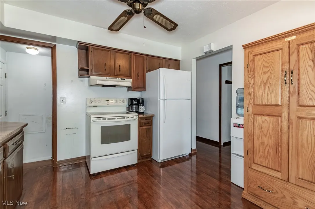 Kitchen with white appliances, dark wood-style floors, a ceiling fan, under cabinet range hood, and brown cabinets
