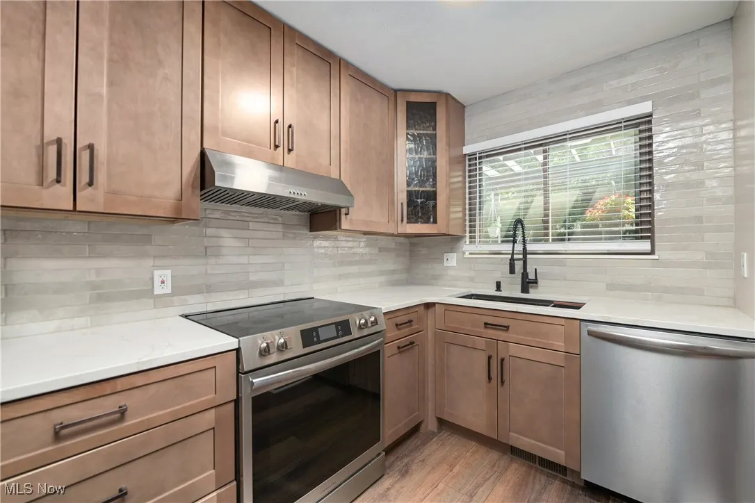 Kitchen with appliances with stainless steel finishes, under cabinet range hood, light stone counters, and decorative backsplash