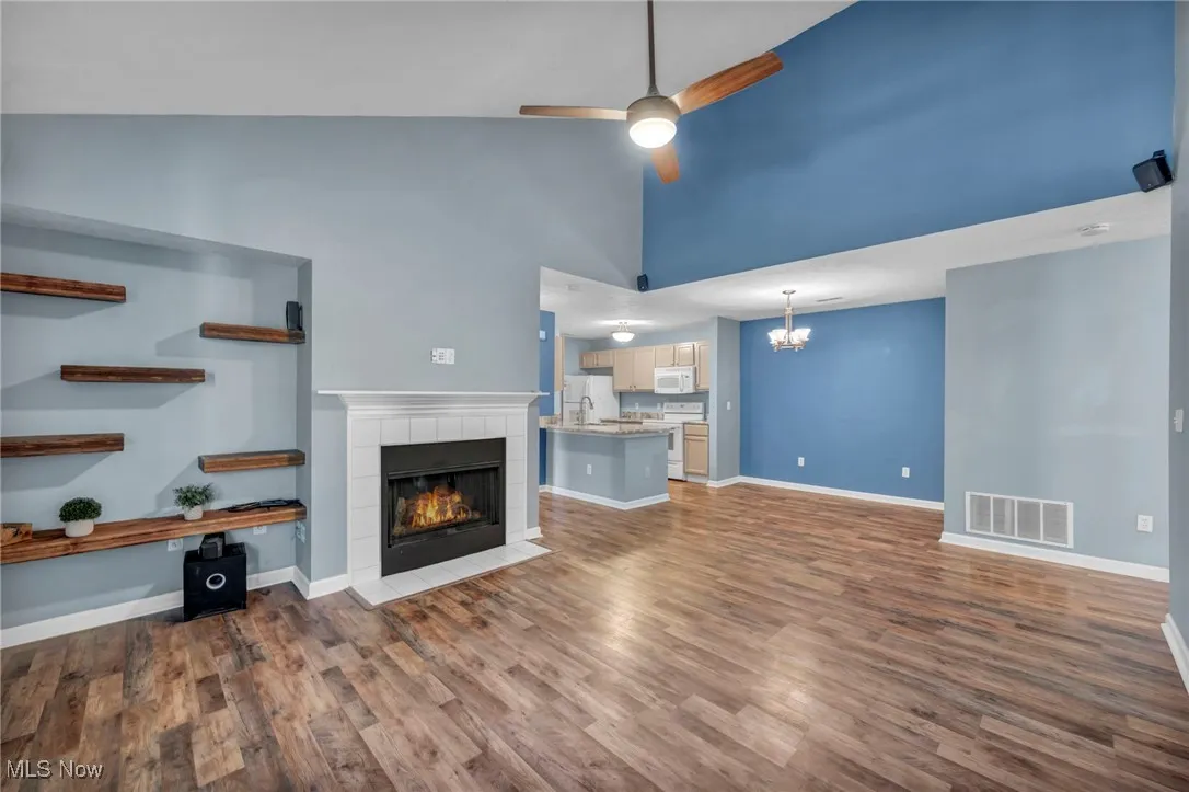 Unfurnished living room featuring high vaulted ceiling, a tiled fireplace, dark wood-style flooring, a ceiling fan, and a chandelier