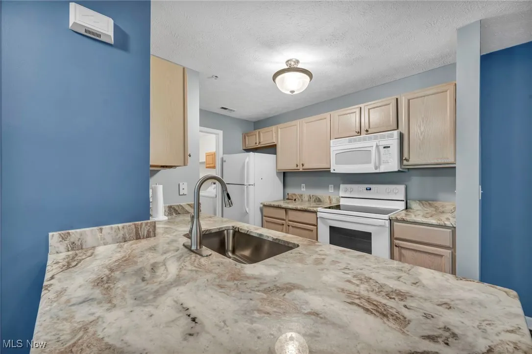 Kitchen featuring light brown cabinetry, white appliances, light stone countertops, and a textured ceiling