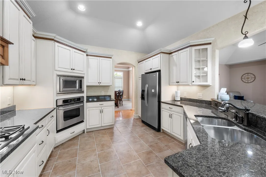 Kitchen with arched walkways, appliances with stainless steel finishes, light tile patterned floors, white cabinetry, and dark stone counters