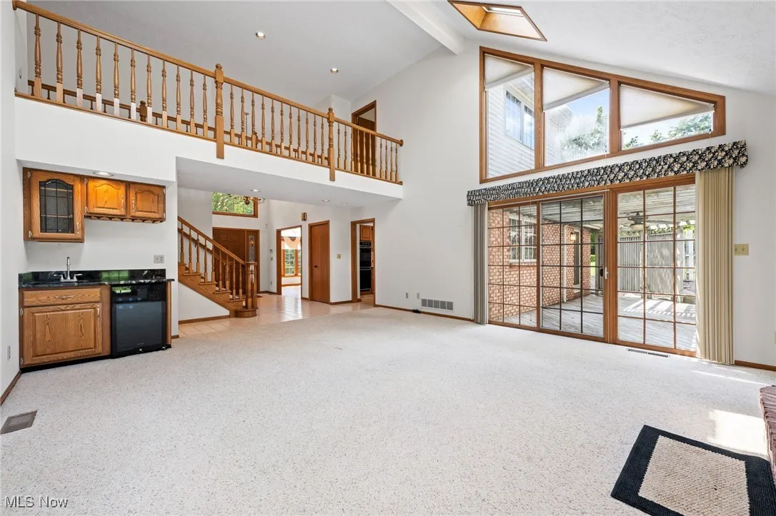 Unfurnished living room featuring stairs, light carpet, beam ceiling, high vaulted ceiling, and bar with sink