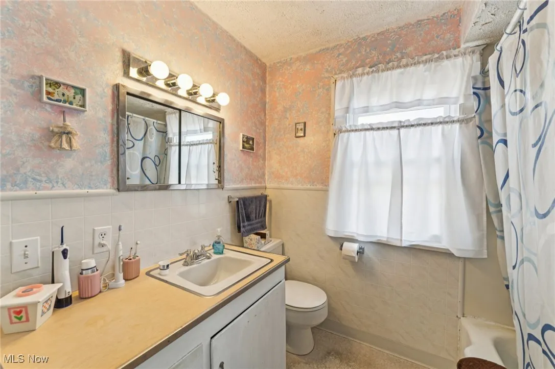 Bathroom with wallpapered walls, a textured ceiling, vanity, shower / tub combo with curtain, and tile walls